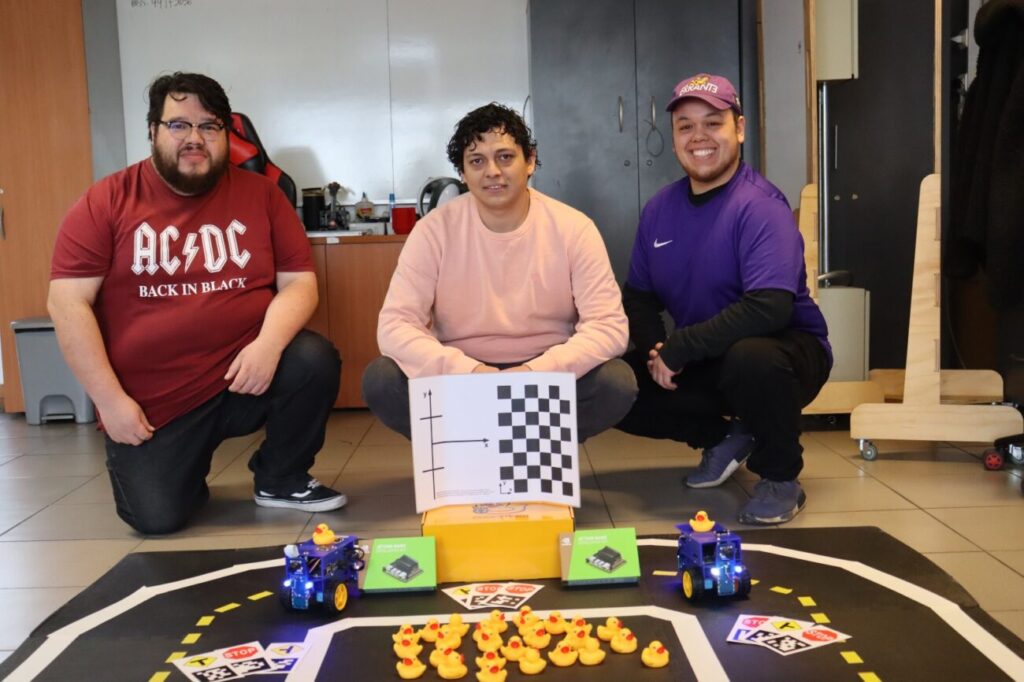 Felix donoso and two colleagues kneeling behind a duckietown setting (duckietown, duckiebots, traffic signs)