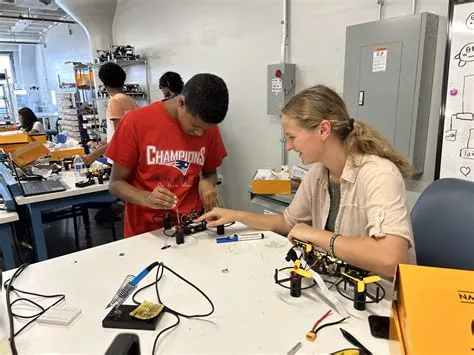 Two students in a workshop build small robot kits at a lab table, with a soldering iron, wires, and tools nearby.